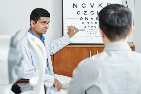 A doctor in a white coat is pointing at a letter chart. Another man seen from the back is reading the letter chart.