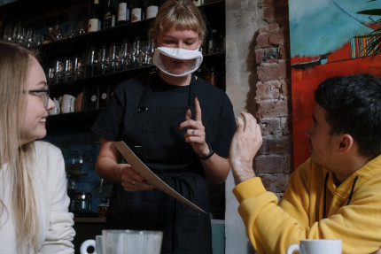Set in a cafe, two people are ordering from the menu. A third person, the waitress is wearing a transparent mask to allow lip reading. People are aslo making handsigns.