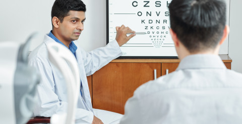 A doctor in a white coat is pointing at a letter chart. Another man seen from the back is reading the letter chart.